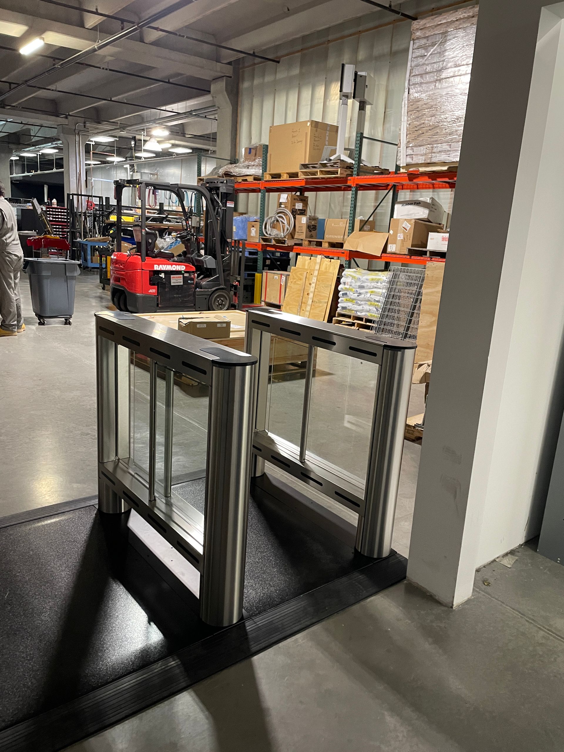 Two silver and glass security turnstiles on a black mat in a warehouse, with a forklift and shelves in the background.