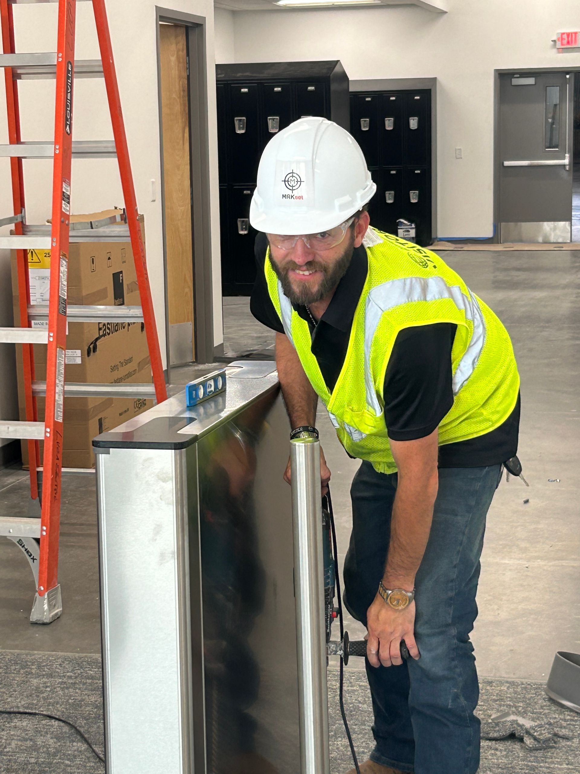 Construction worker in hard hat and safety vest using a tool on a silver cabinet.
