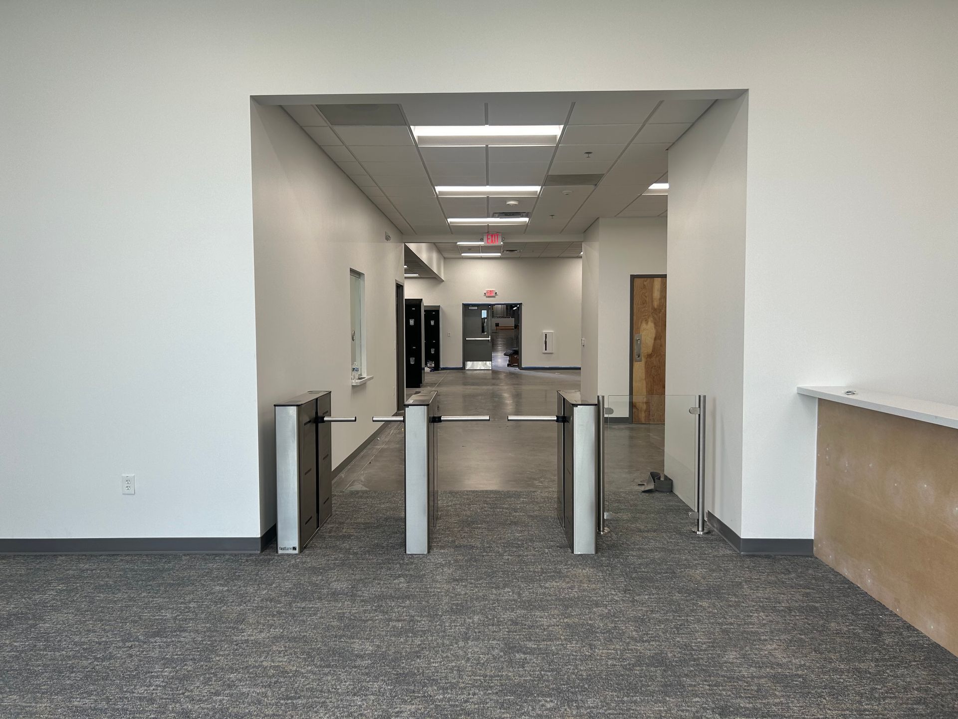 Hallway entrance with three turnstiles, carpet floor, white walls, and an exit sign visible down the hall.