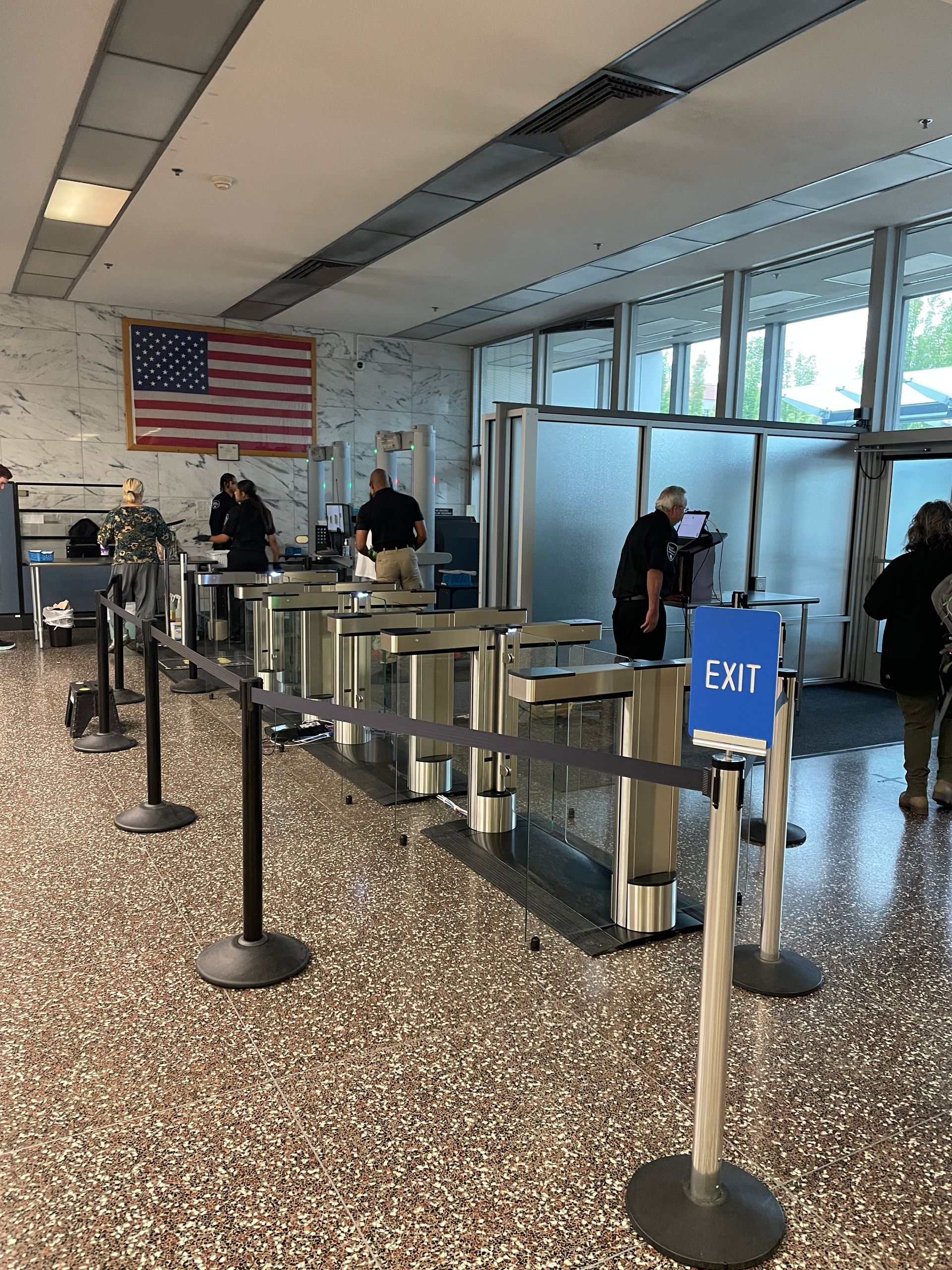 Airport security checkpoint: People passing through turnstiles, U.S. flag on the wall.