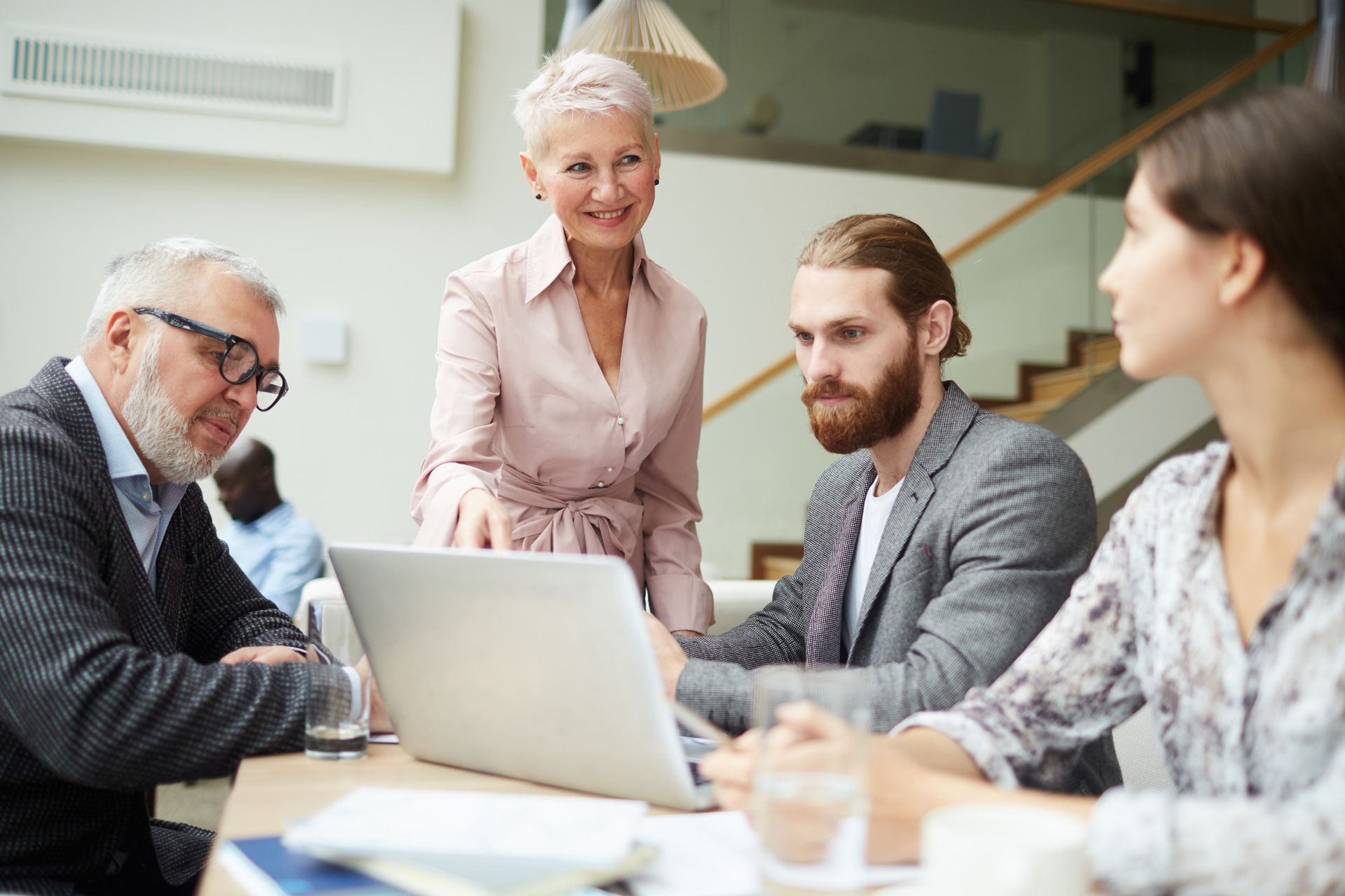Four colleagues collaborating around a laptop in a modern office, with one pointing at the screen.