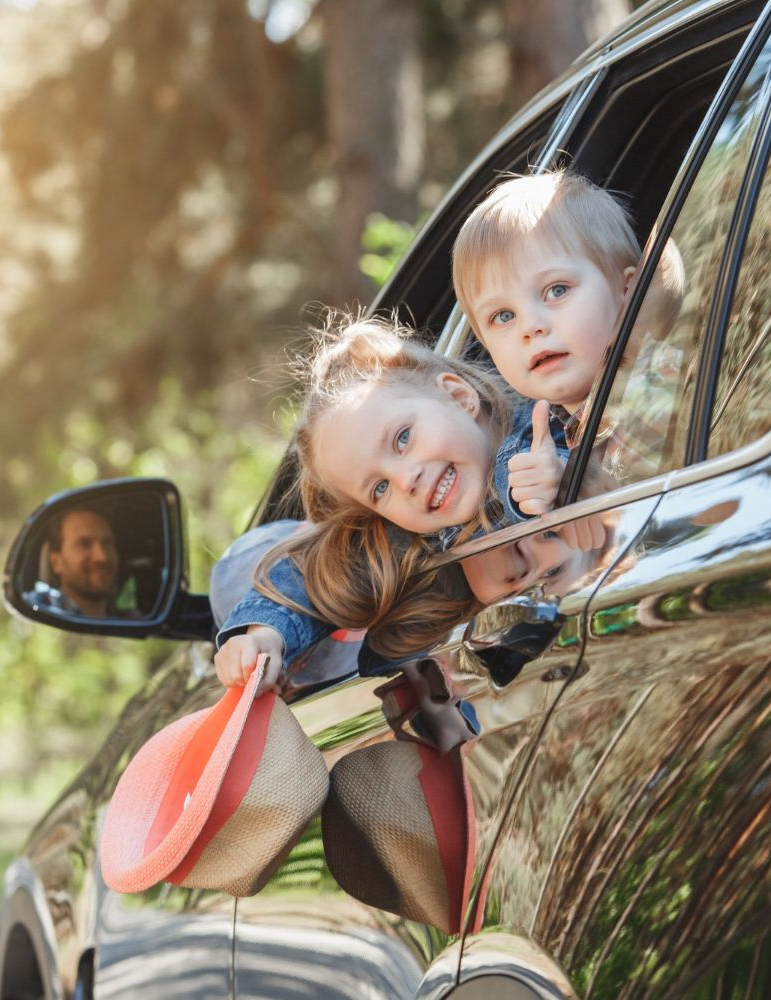 Two children in car window, smiling and looking out; person reflected in side mirror.
