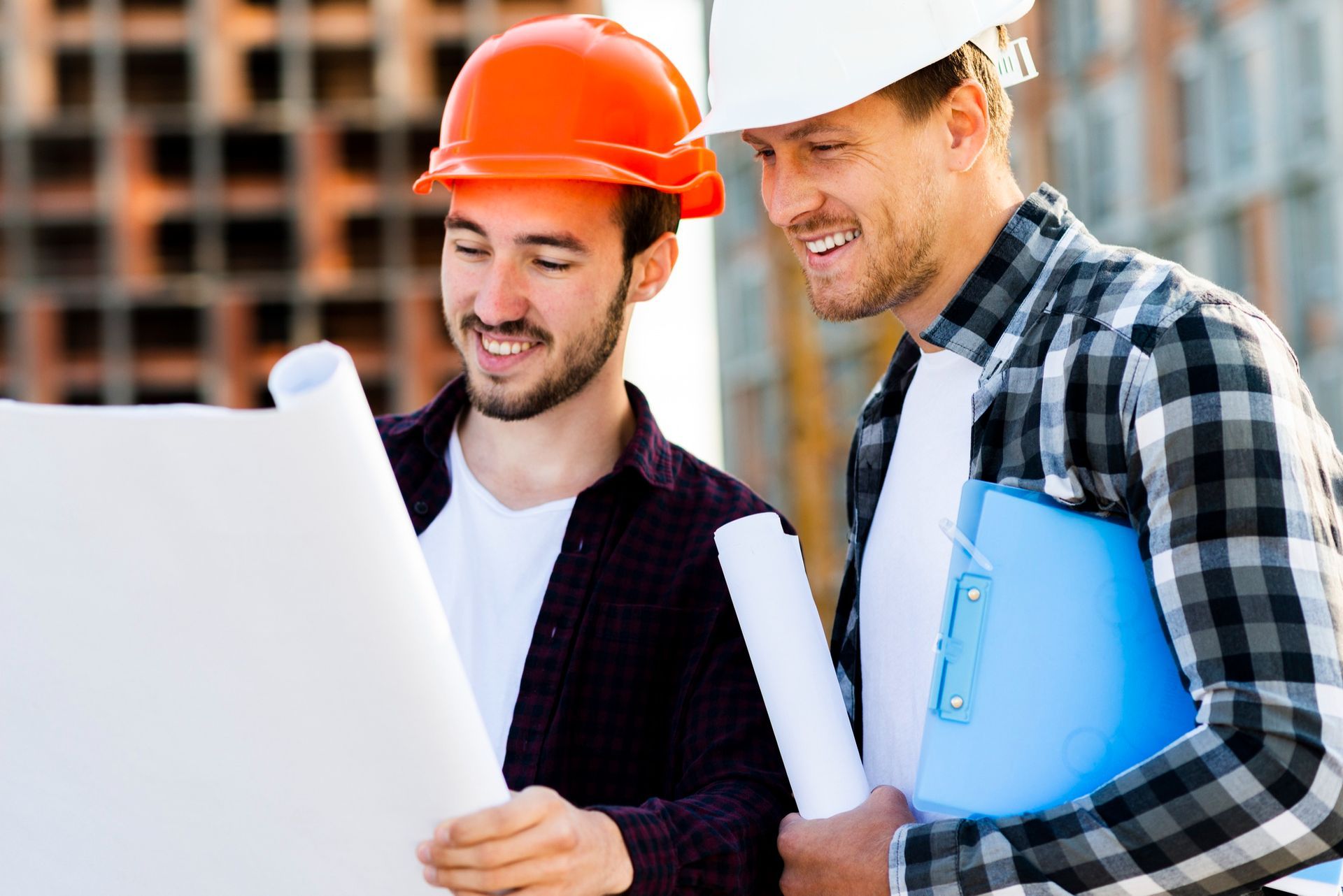 Two construction workers in hard hats examine blueprints, smiling, on a construction site.