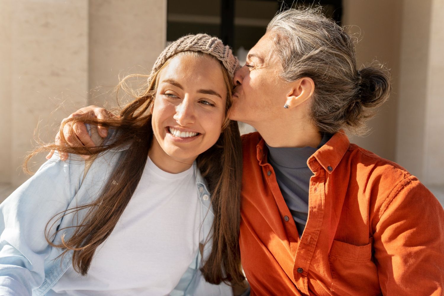 Person kissing the cheek of another person outdoors. One is smiling, wearing a hat, the other has gray hair.
