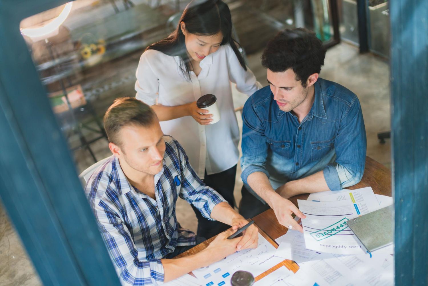 Three people at a table, reviewing papers. One points at data, while another holds a phone. A third person holds a coffee cup.