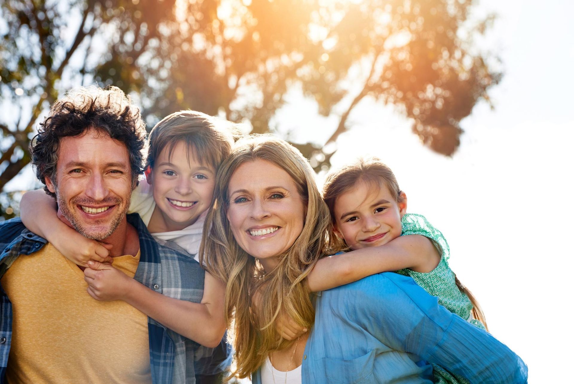 Family smiles at the camera outdoors, two children on parents’ backs, sunlit, happy expressions.
