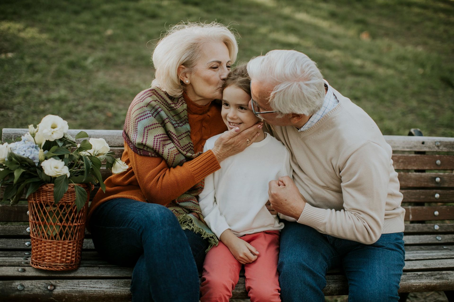 Grandparents kissing granddaughter on the cheeks, sitting on a bench with flowers.