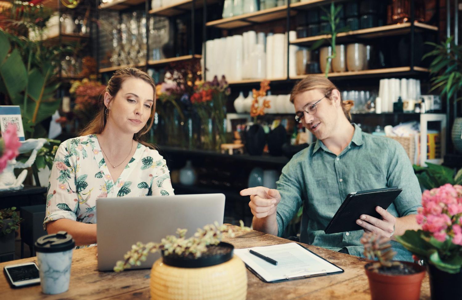 Woman using laptop, man pointing at tablet, seated at a table in a floral shop.