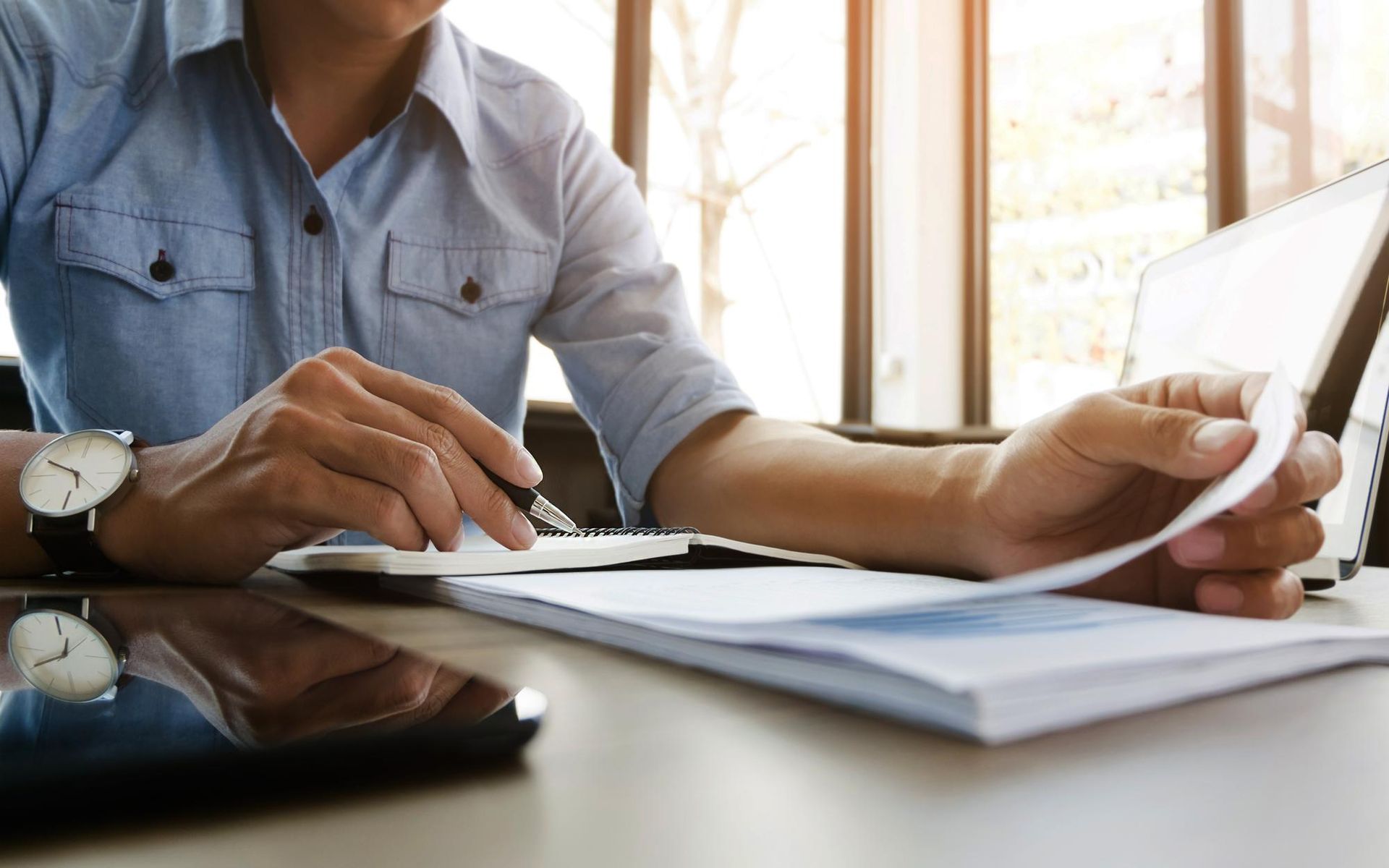 Person in a blue shirt reviews papers at a desk with a laptop, phone, and watch in a sunlit setting.