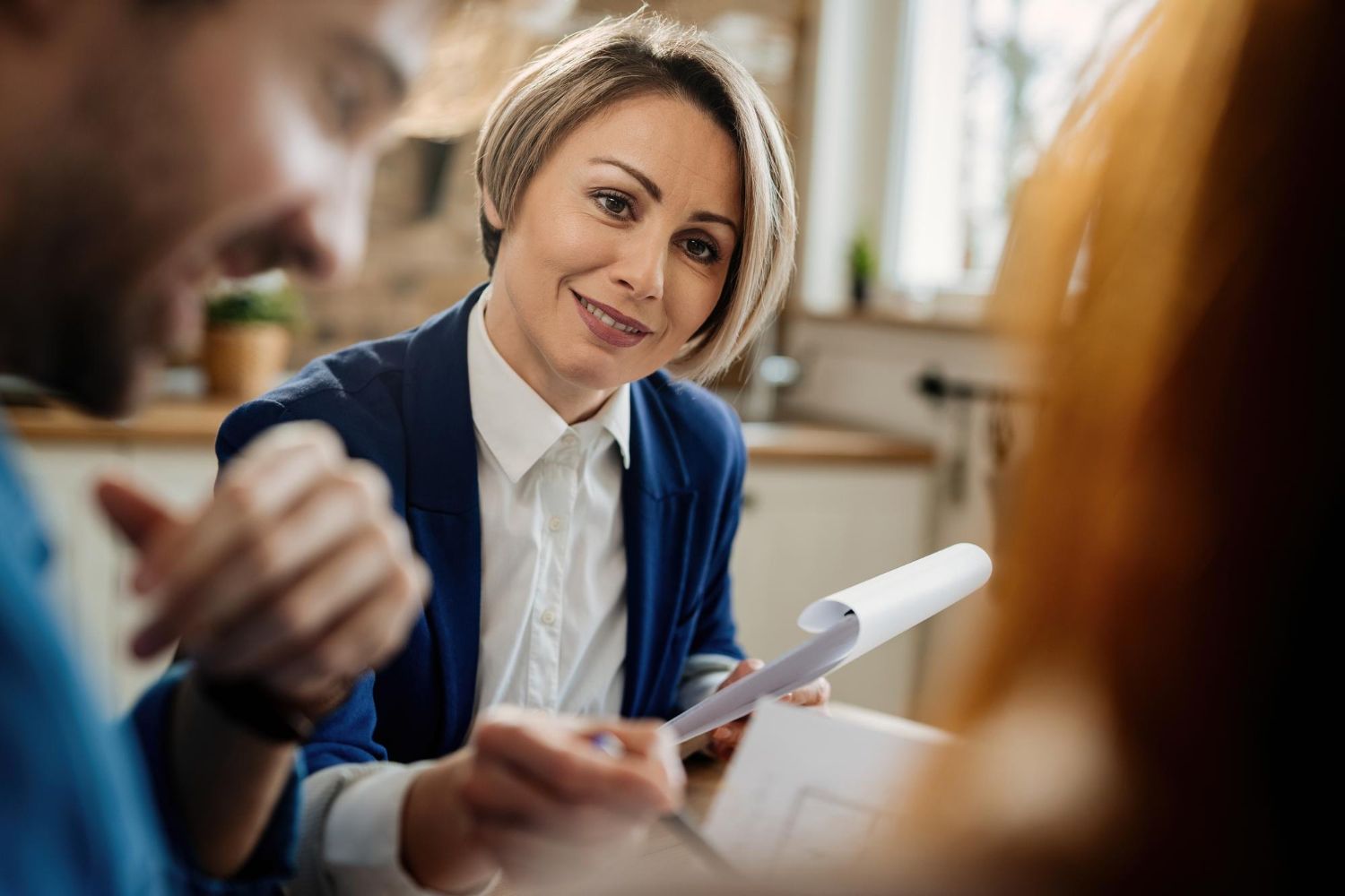Woman in a blue blazer smiles while holding papers and talking to a couple in a room.