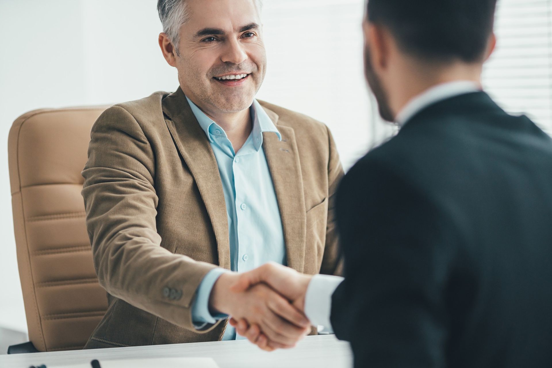 Man in tan jacket shaking hands with another in a black suit, smiling, in an office.