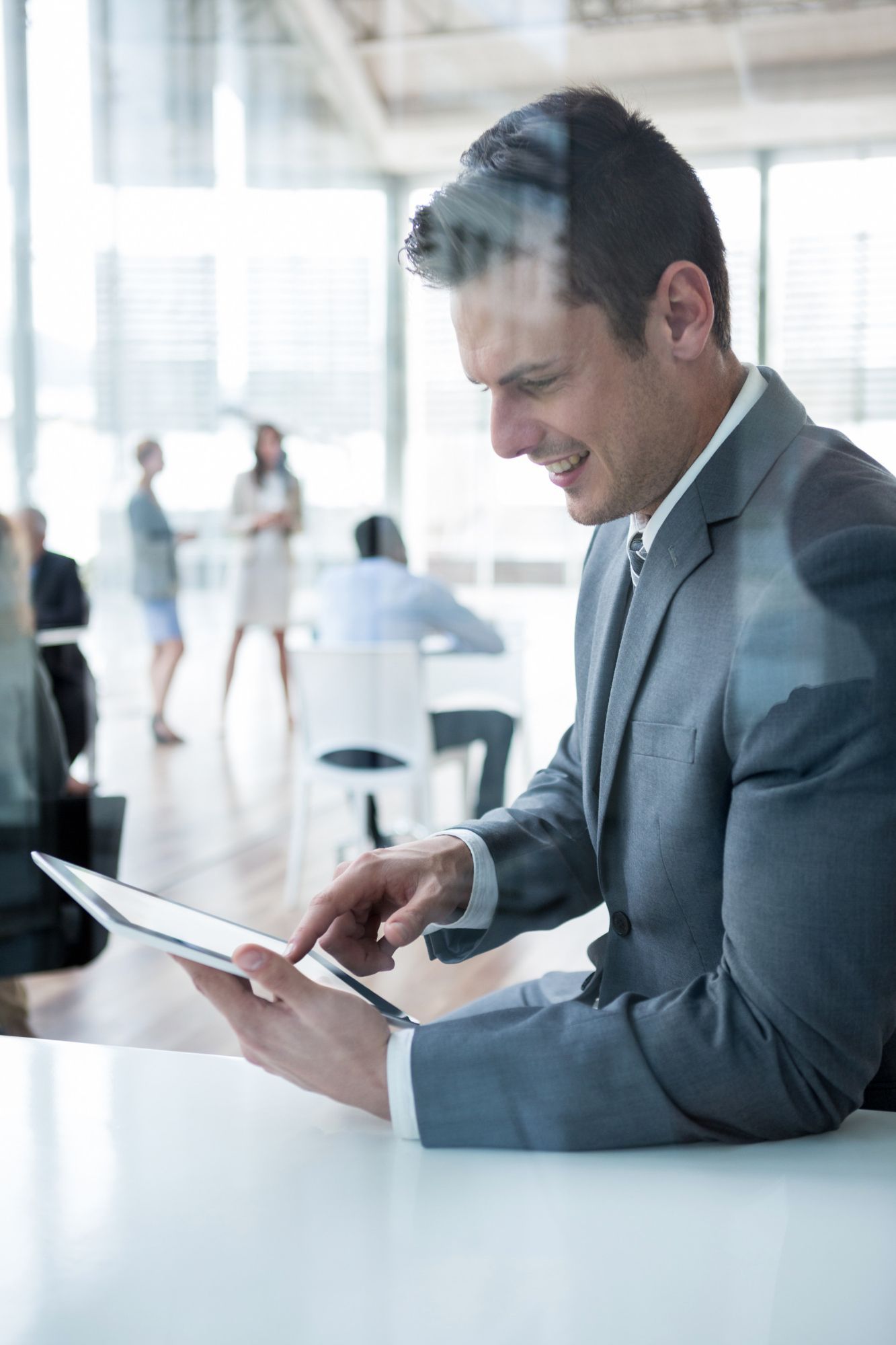 Man in suit using a tablet in a modern office, smiling. Other people visible in the background.