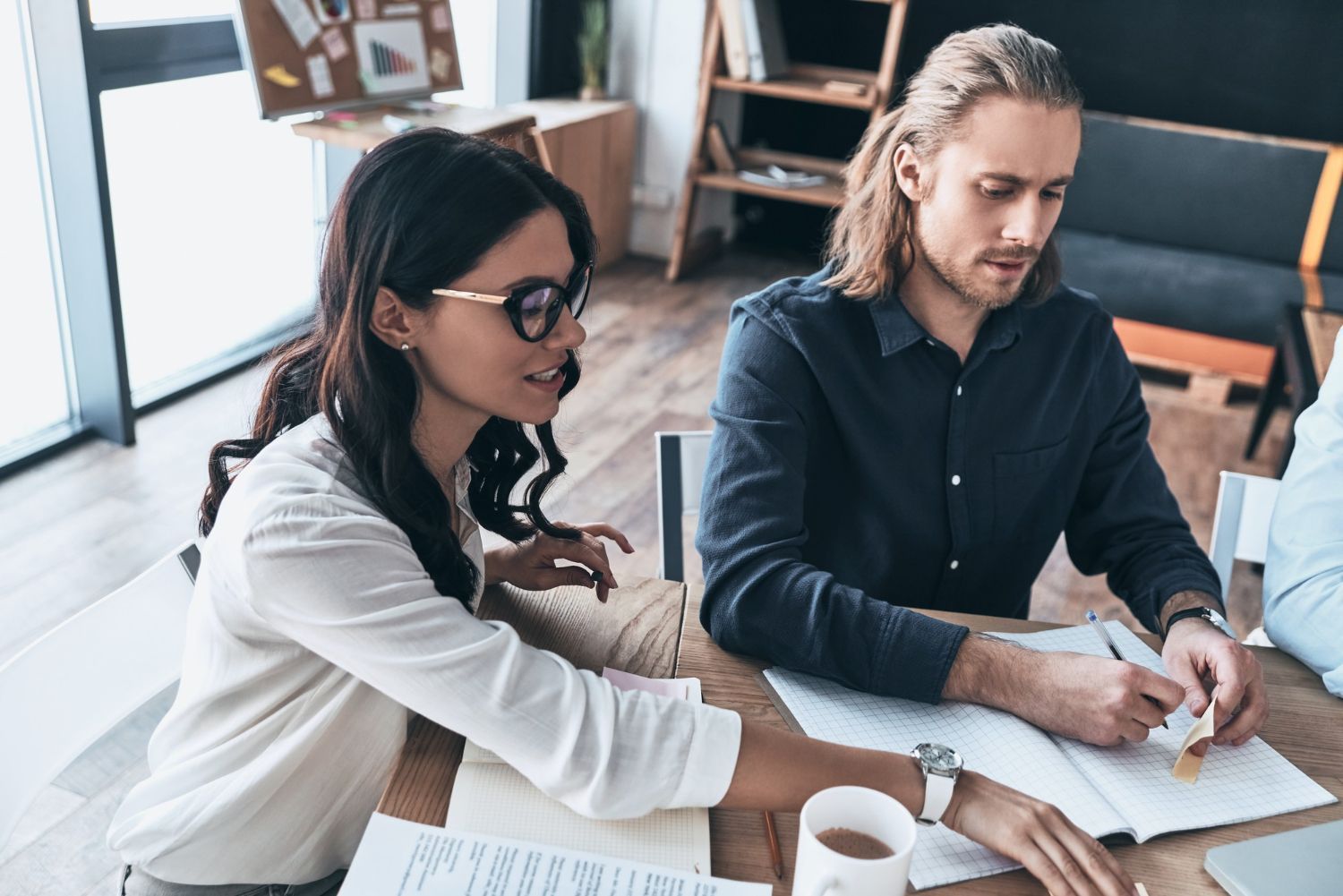 Woman in glasses points at paperwork while man with long hair writes, in an office setting.
