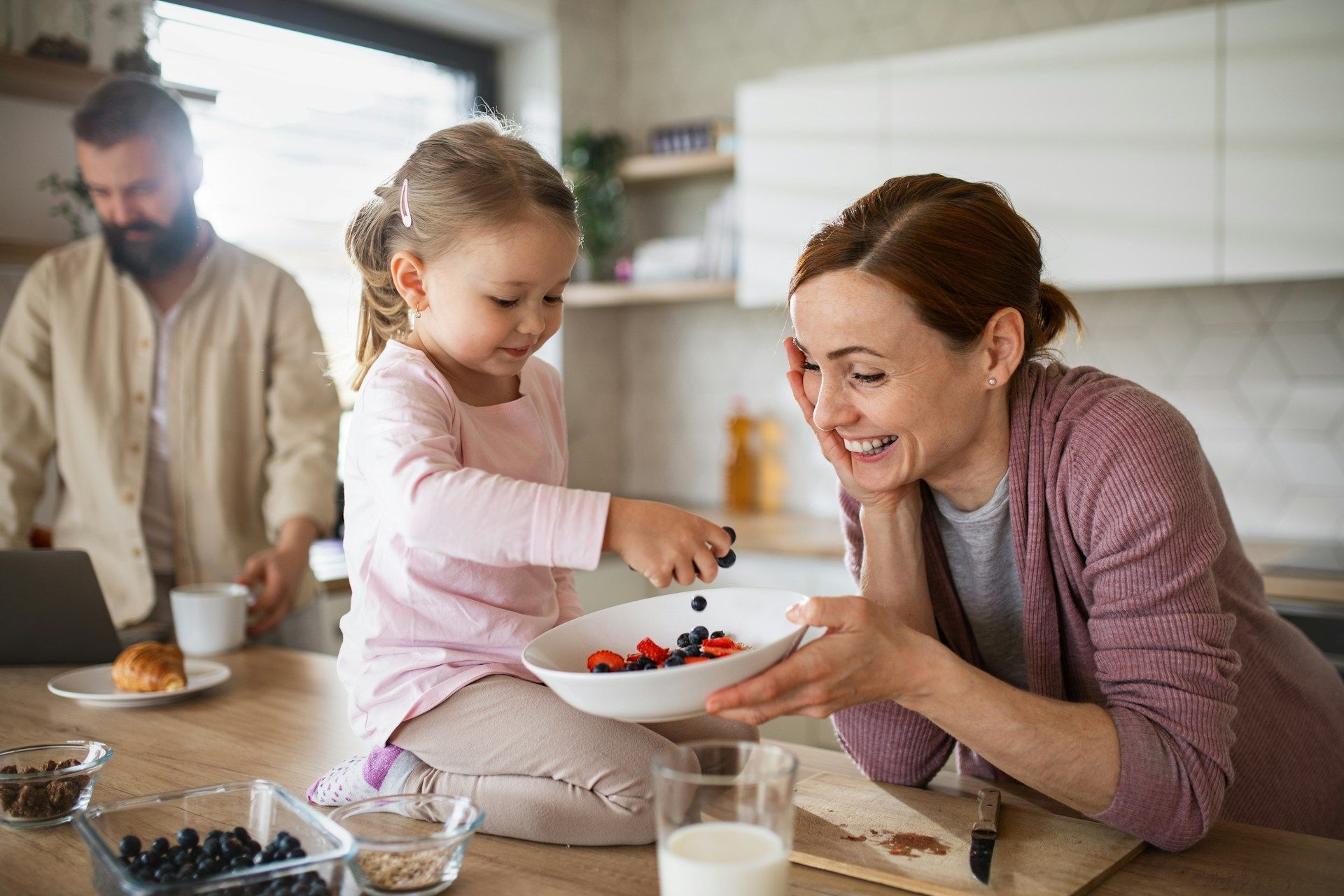 Woman and child prepare fruit bowl in a kitchen, man in background.