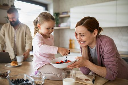 Woman and child prepare fruit bowl in a kitchen, man in background.