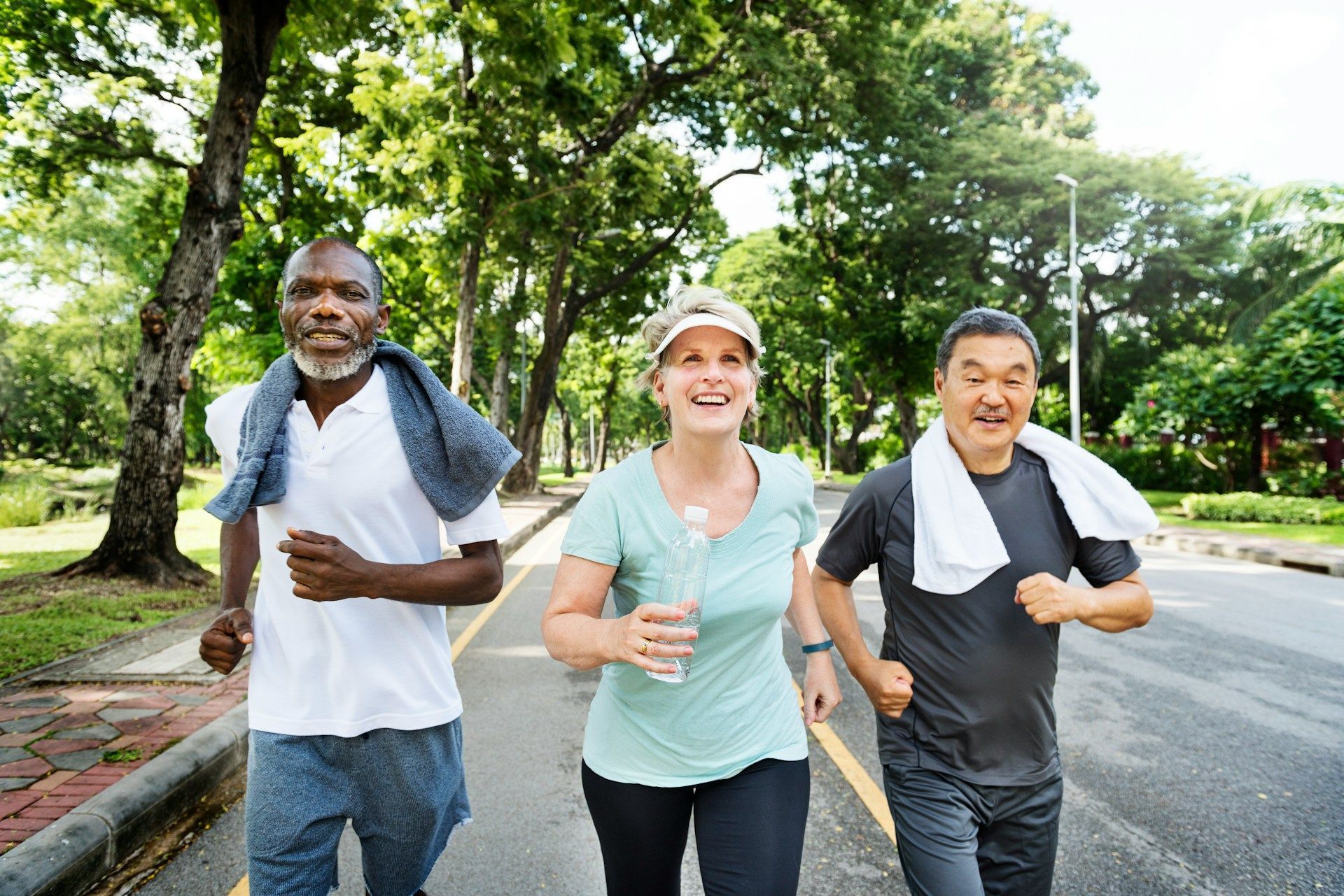 Three people jogging on a paved path in a park; sunny, trees in background.