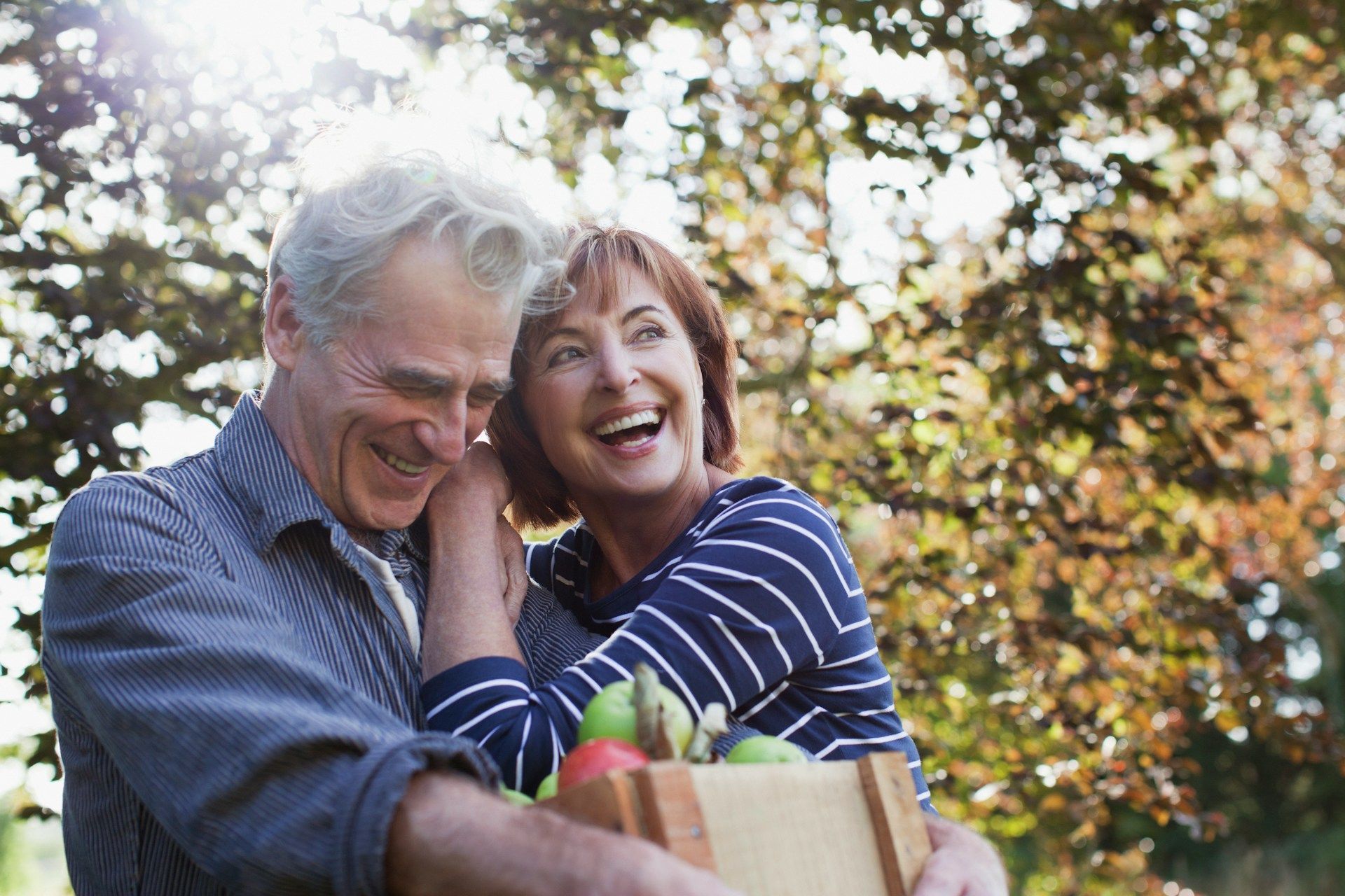 Couple laughing, carrying a wooden crate of fruit in a sunny outdoor setting.