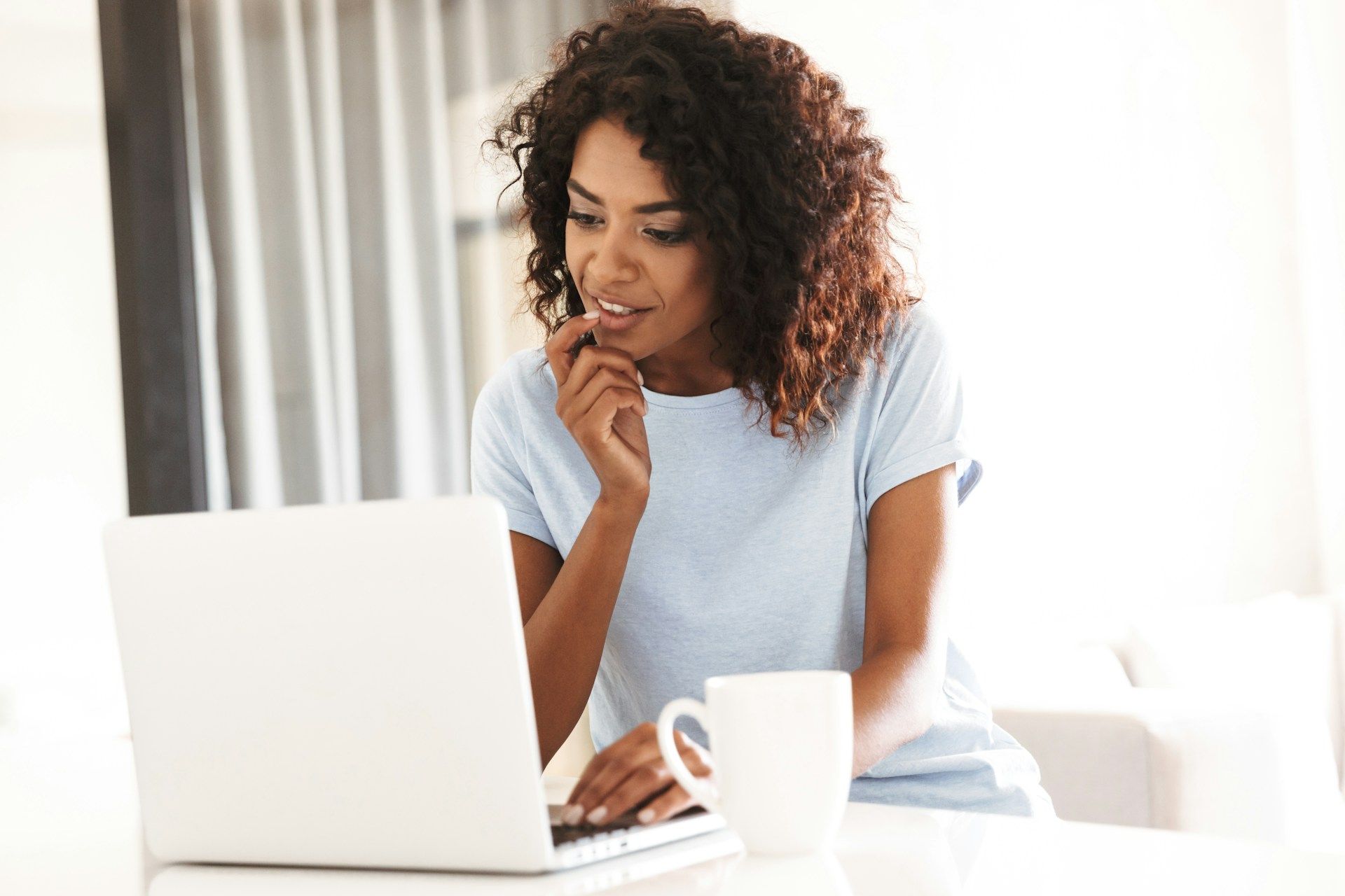 Woman with curly hair works on a laptop, hand to chin, with a mug on a white counter.