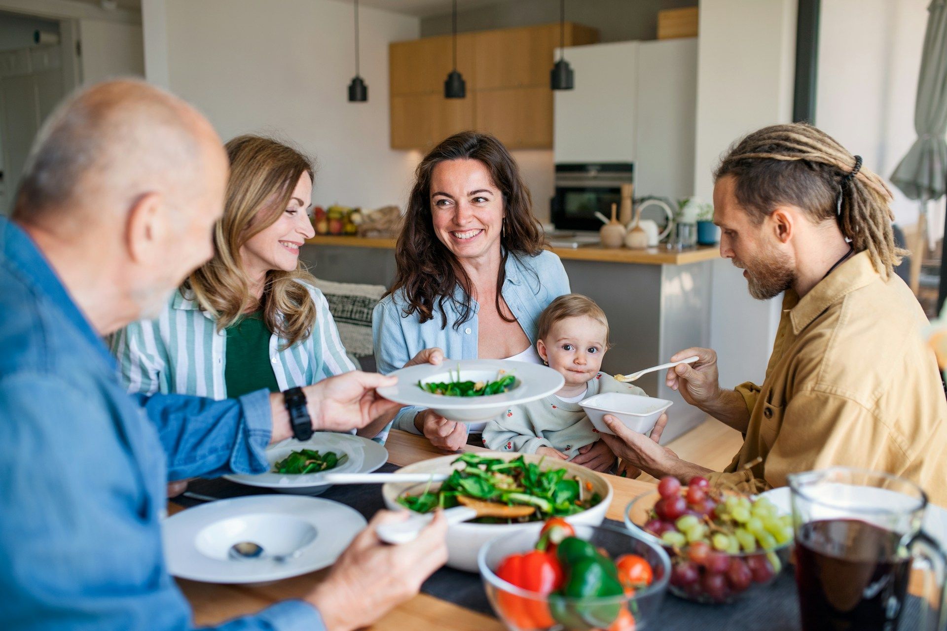 Family at a table, eating. Adult children, baby, and parents enjoy a meal together in a bright kitchen.
