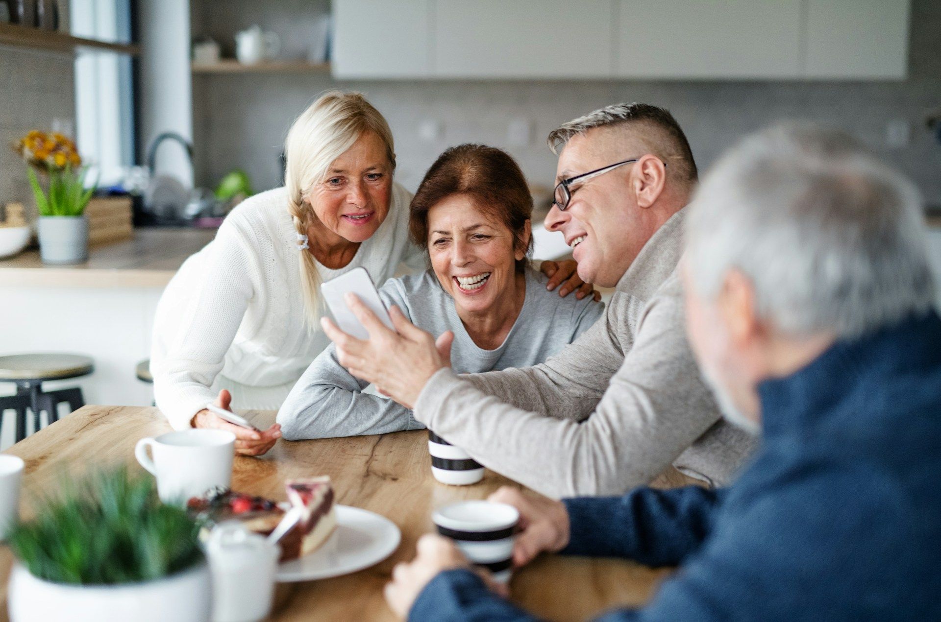 Four people looking at a phone, smiling around a kitchen table with coffee cups and cake.