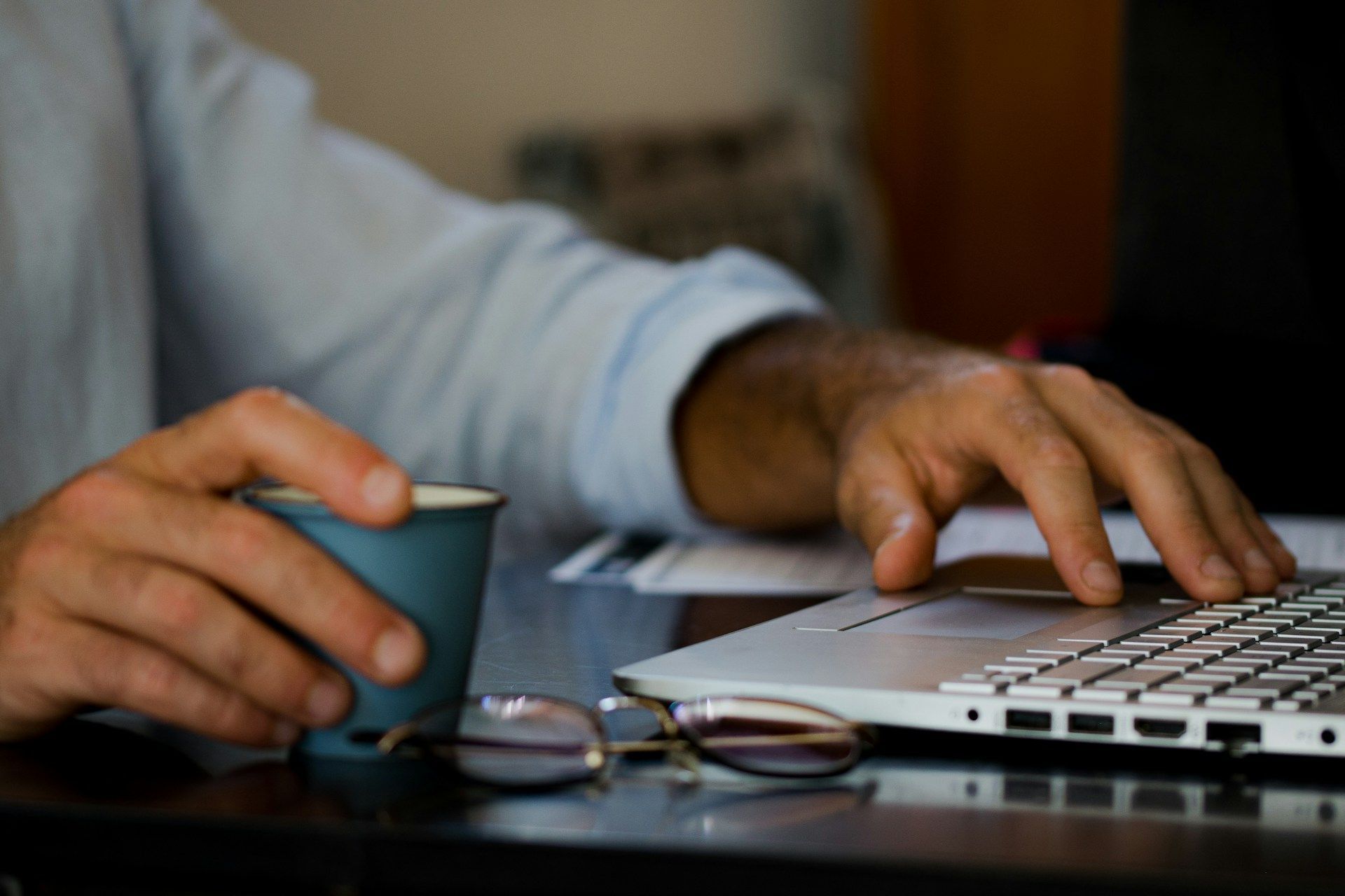 Person using a laptop, holding a mug. Glasses sit on the table.