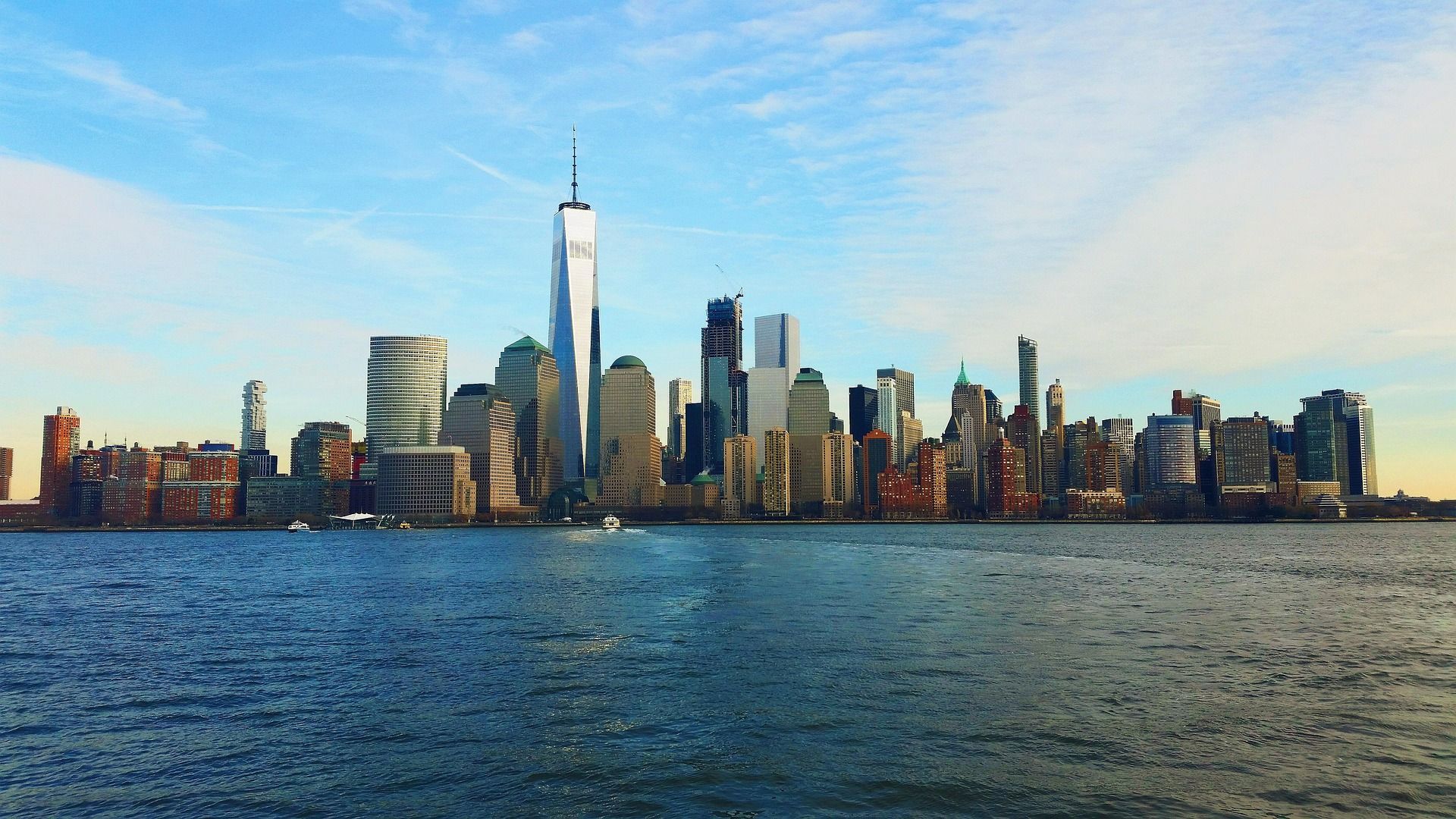 New York City skyline on a sunny day, seen across blue water. One World Trade Center stands tall.