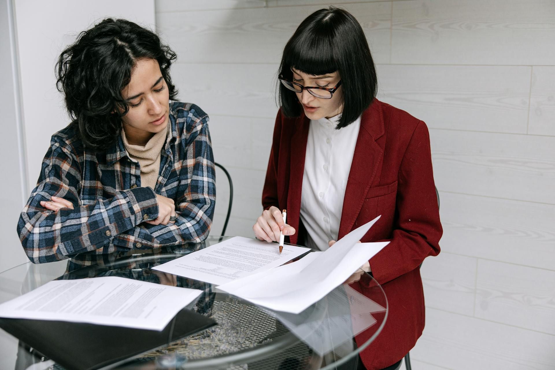 Two people reviewing documents at a table. One points while the other looks on with crossed arms.