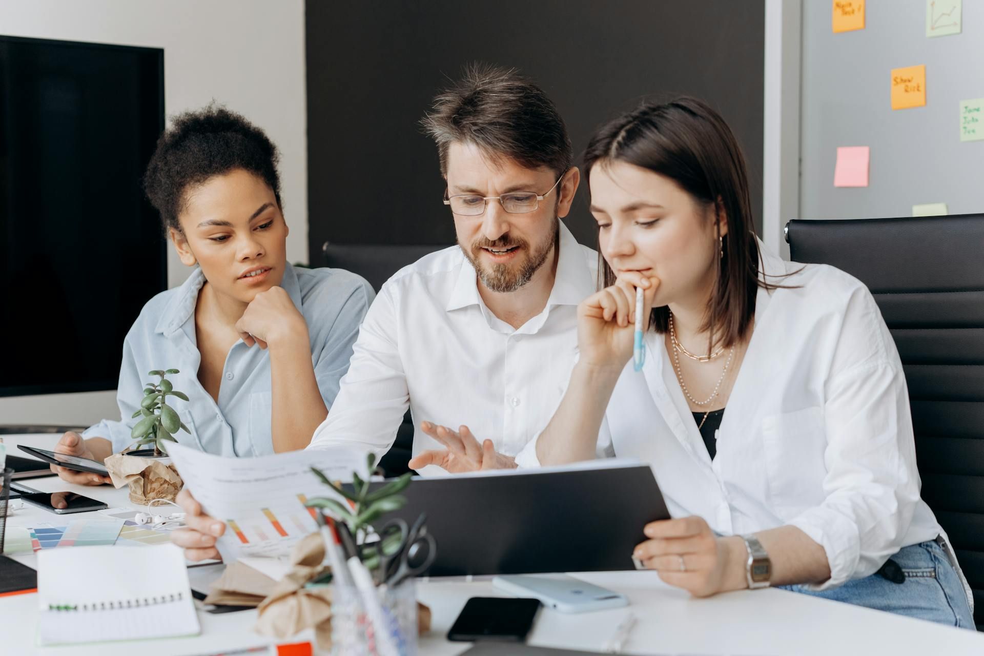 Three people reviewing documents at a desk in an office. They appear focused and are pointing at papers and a tablet.