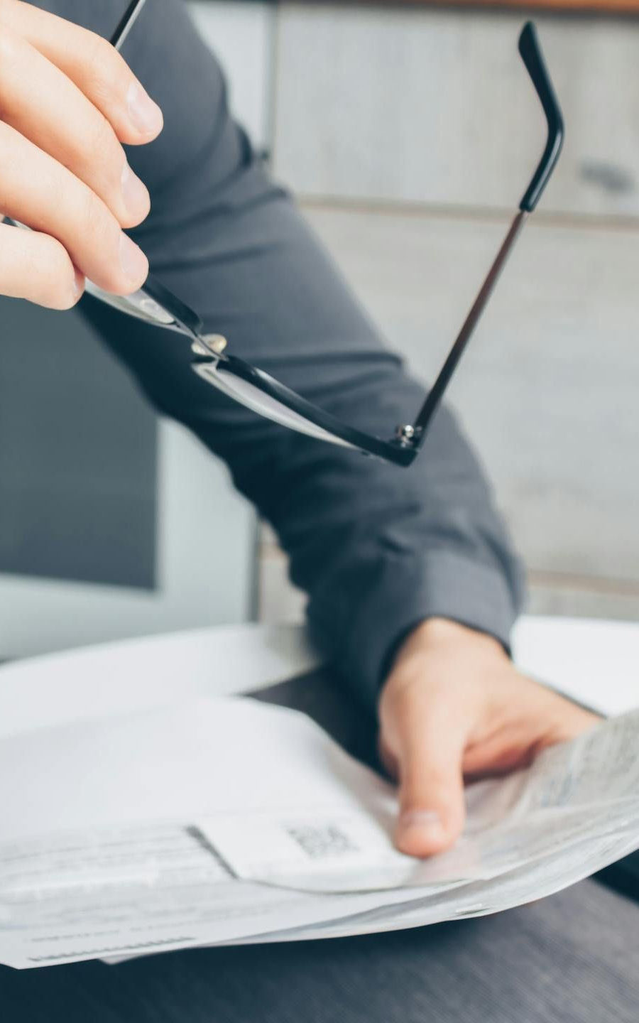 Person holding glasses, looking at a newspaper. White paper and gray long-sleeved shirt visible.