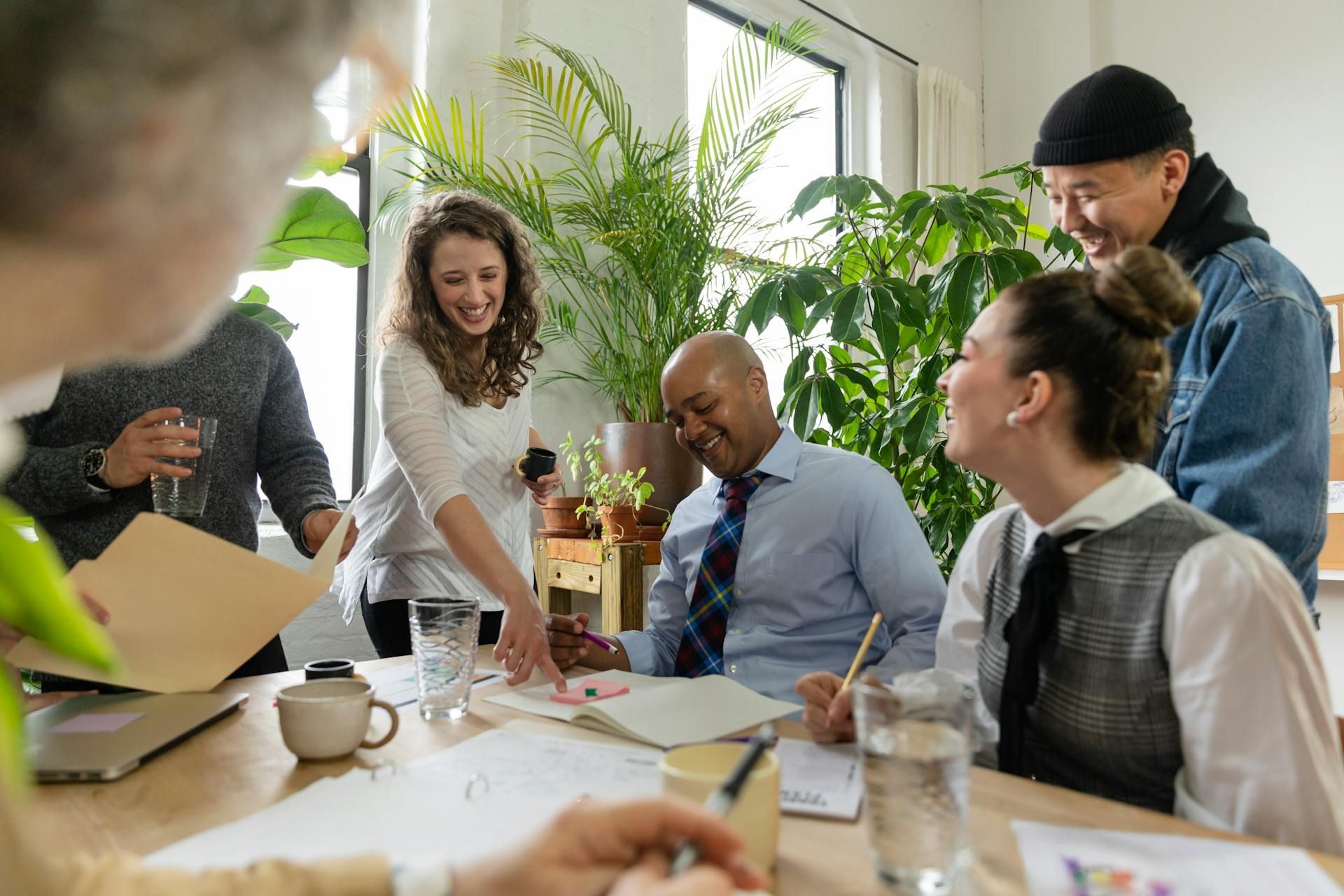 People collaborating around a table with papers, smiling and looking at documents in a bright room with plants.