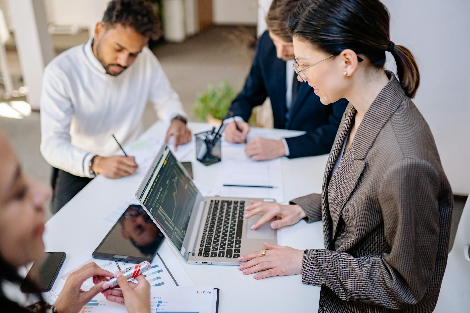 People in business attire working at a table, using laptops and writing, in an office.