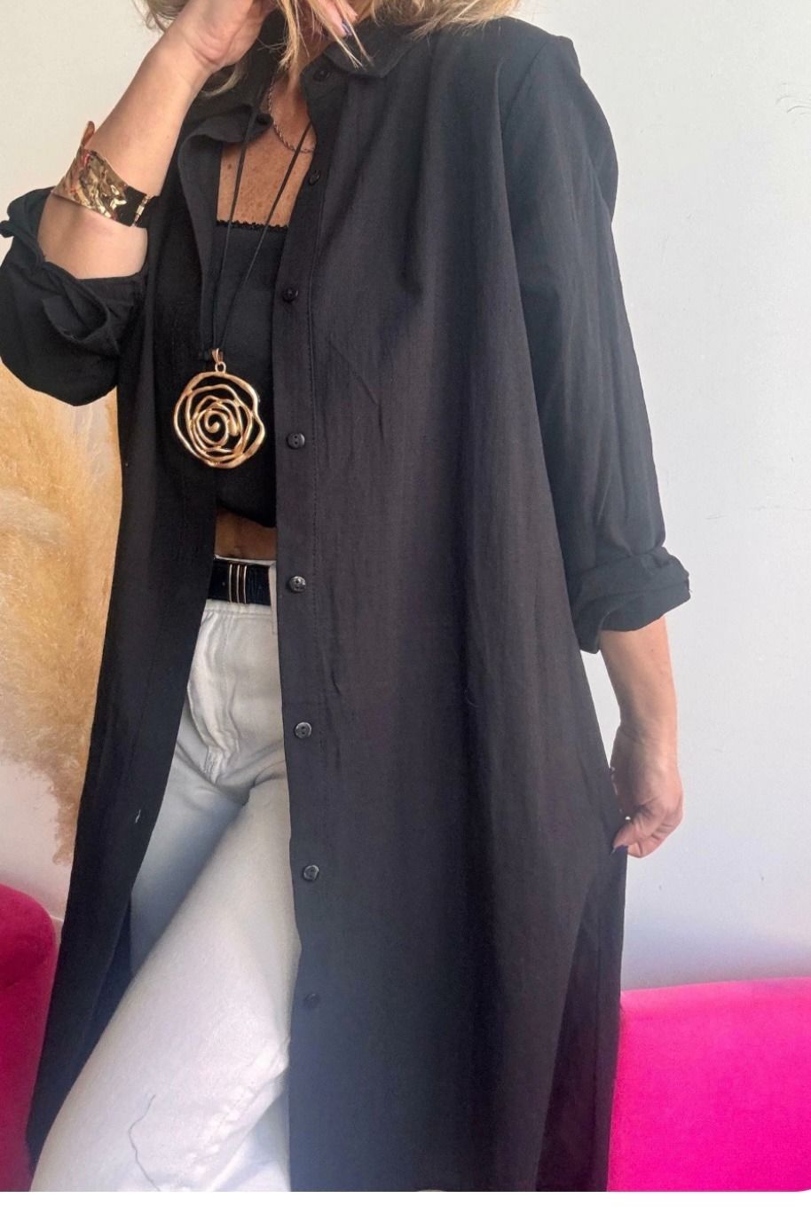 Mujer con camisa negra tipo gabardina sobre top negro y pantalones blancos; brazo levantado, joyas de oro.