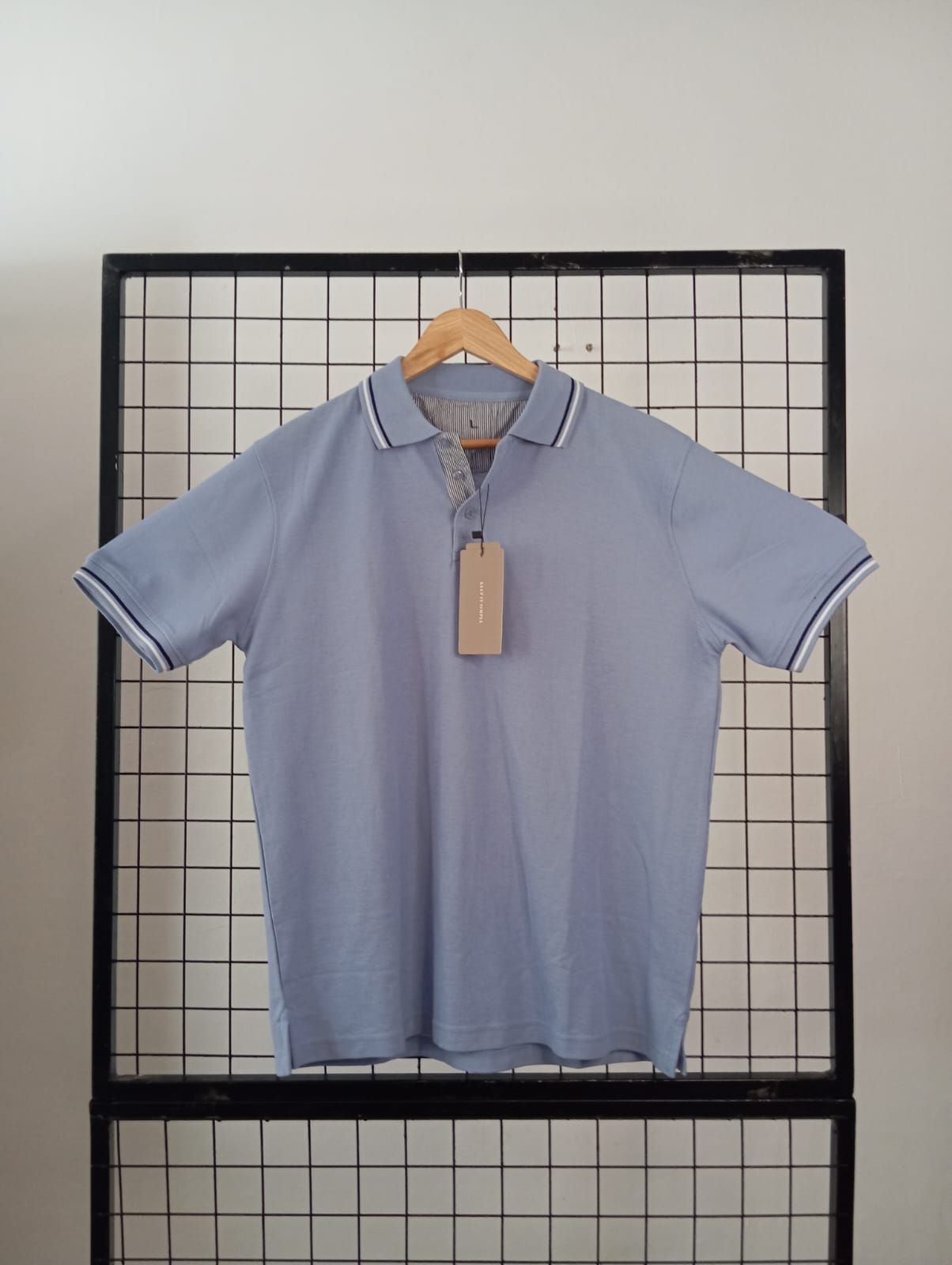 Polo azul claro con ribetes blancos y azul marino, colgado en una percha de madera sobre un fondo cuadriculado.