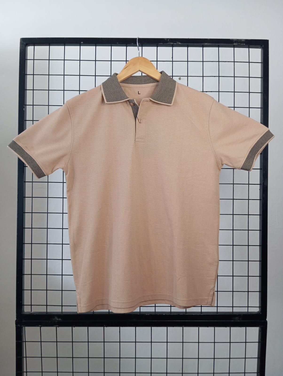 Polo beige con ribete marrón en una percha de madera, expuesto sobre una rejilla metálica negra.