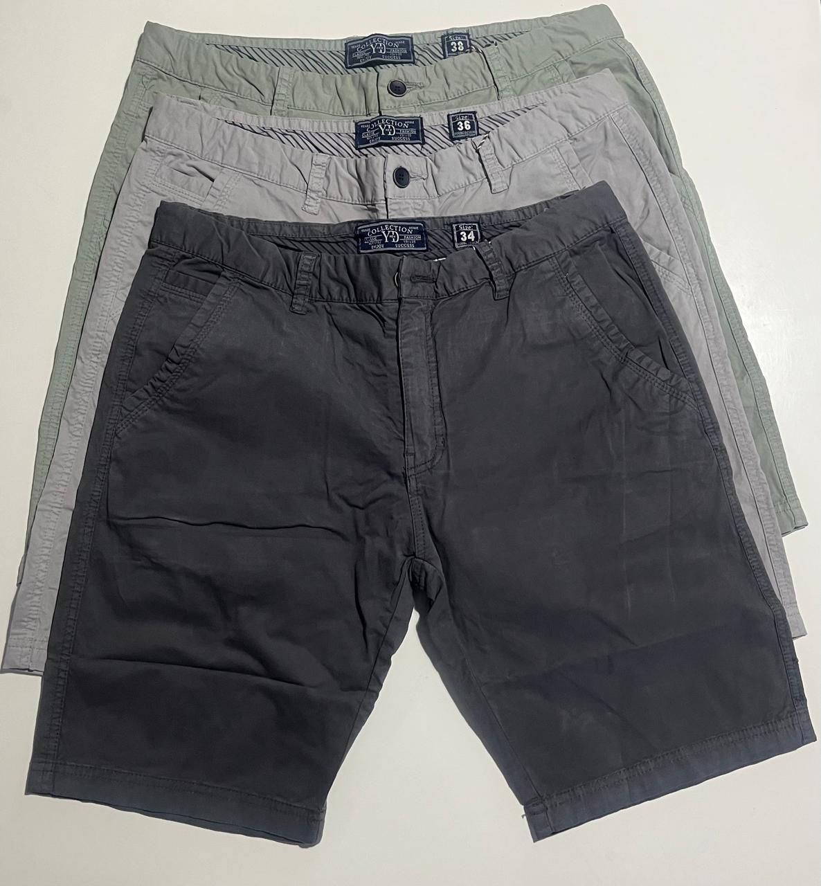 Tres pares de pantalones cortos color caqui en diferentes tonos de verde, gris y negro, apilados sobre una superficie blanca.