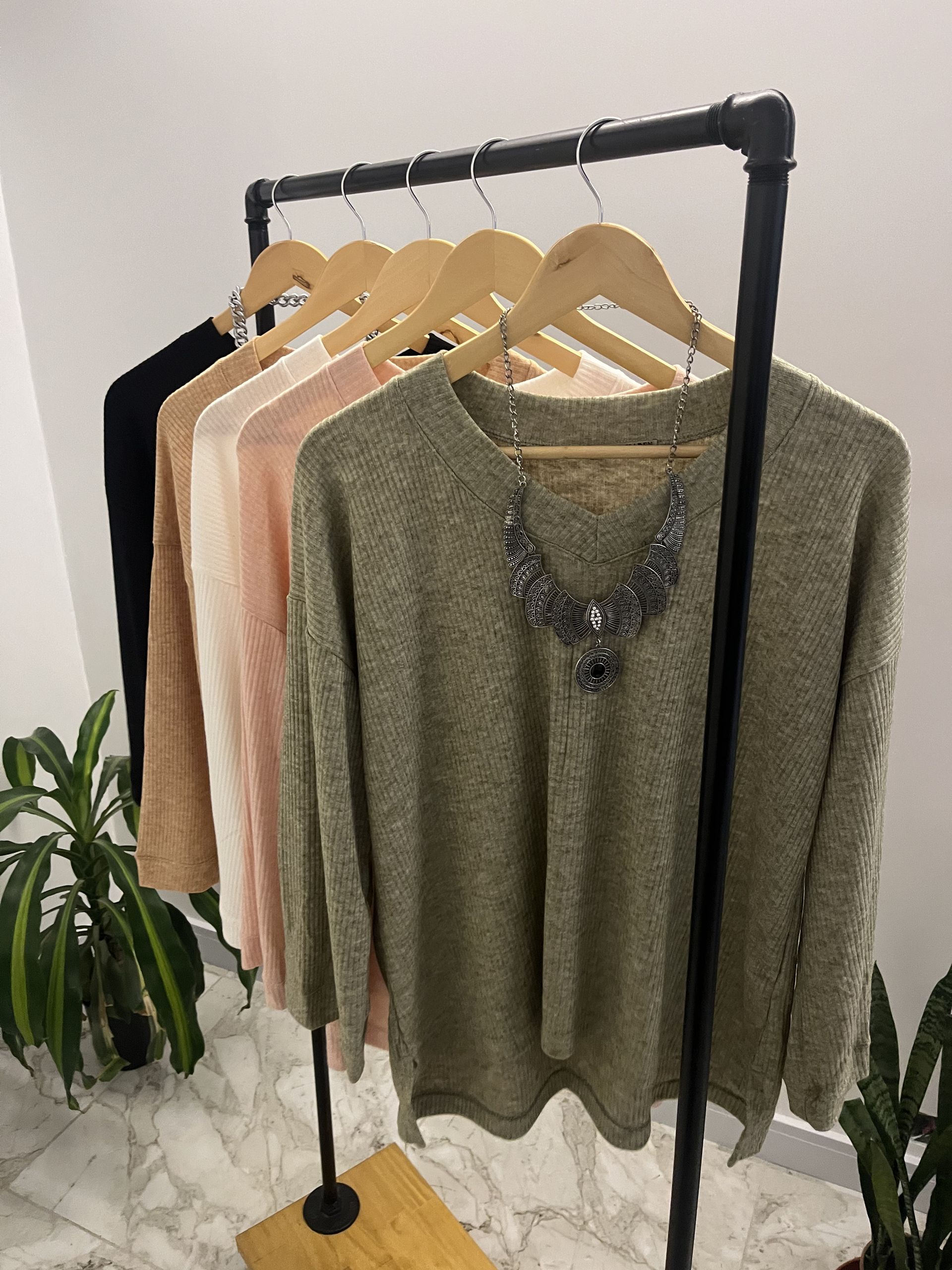 Perchero con cinco suéteres en colores neutros: verde oliva, rosa, beige y negro, colgados en perchas de madera.