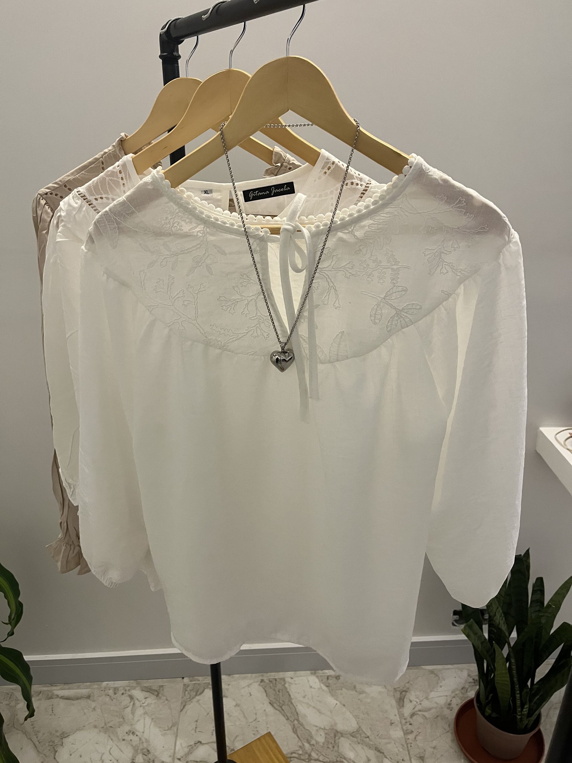 Blusa blanca de encaje en una percha, junto a otras dos blusas; un collar con colgante cuelga de la blusa blanca.