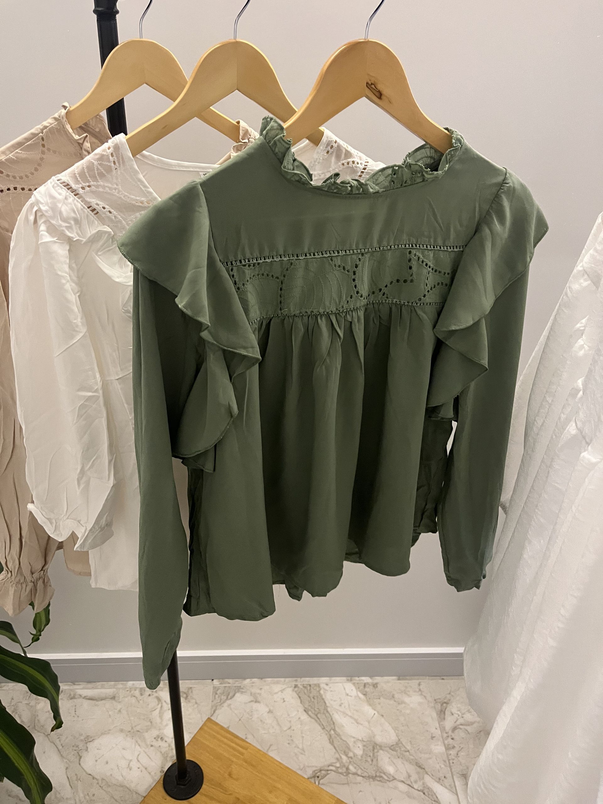 Blusa verde con volantes y detalles de encaje colgada en una percha de madera junto con otras dos blusas.