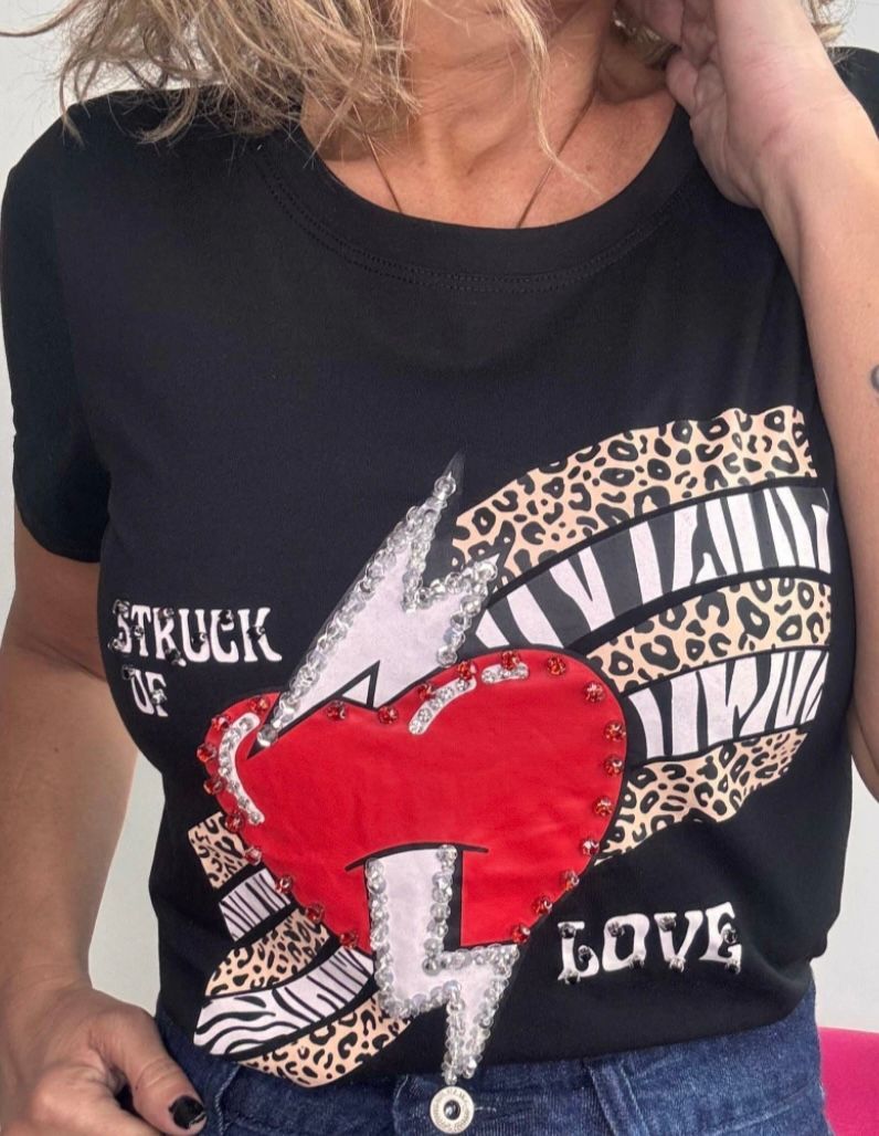 Mujer con camiseta gris estampada con un corazón rojo, un rayo y un animal print; el texto dice “Struck of Love” (Enamorada).