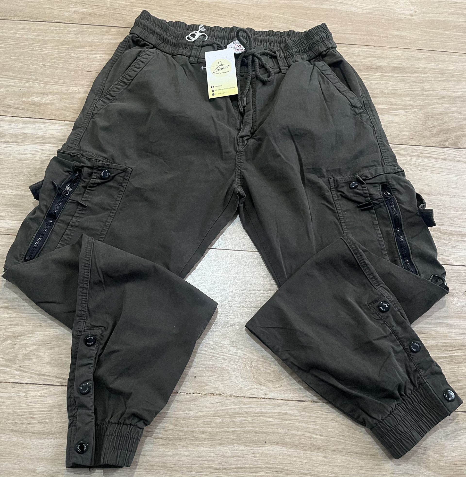 Pantalones cargo de color verde oscuro con múltiples bolsillos y puños abotonados, extendidos sobre madera.
