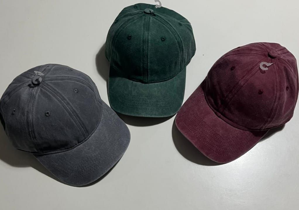 Tres gorras de béisbol de colores: gris, verde y granate, dispuestas sobre una superficie blanca.