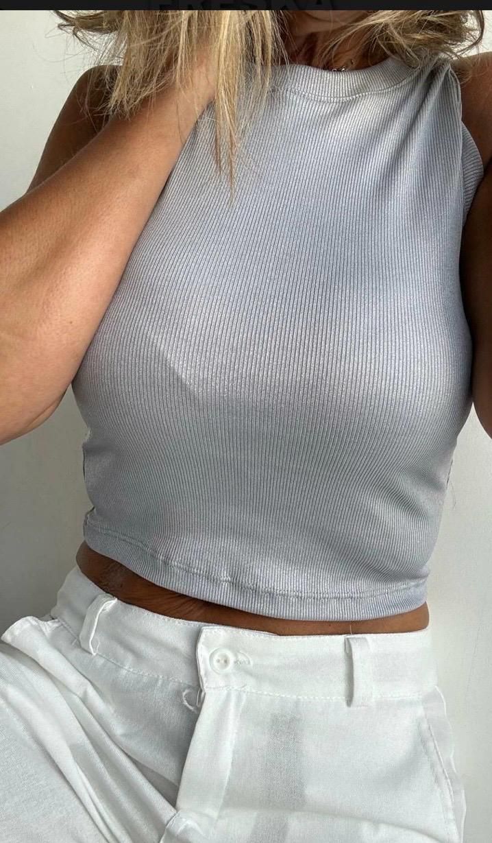 Mujer con top corto de rayas grises y pantalón blanco. Se sujeta el pelo con una mano. Fondo claro.