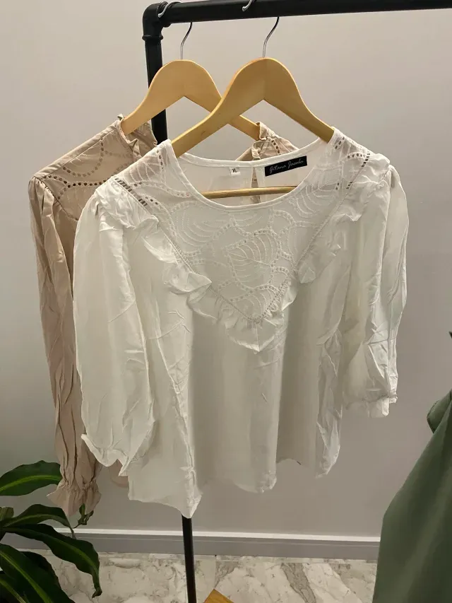Blusa blanca de ojales con volantes y mangas tres cuartos en percha de madera, junto a una blusa beige.
