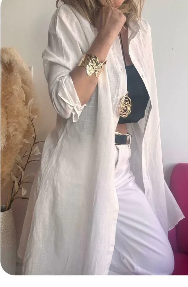 Mujer con camisa negra tipo gabardina sobre top negro y pantalones blancos; brazo levantado, joyas de oro.