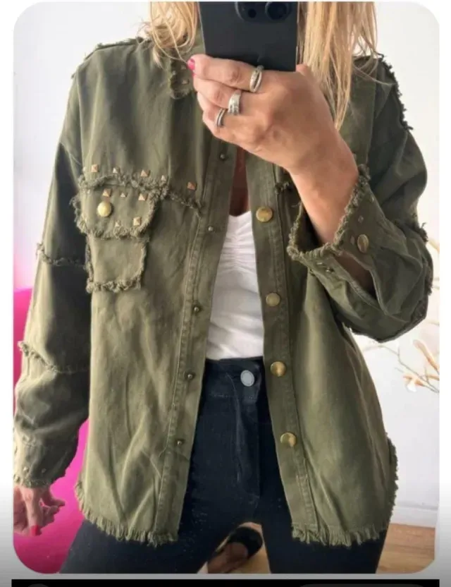 Mujer que lleva una chaqueta vaquera negra con botones y tachuelas doradas sobre una blusa blanca y pantalones negros.