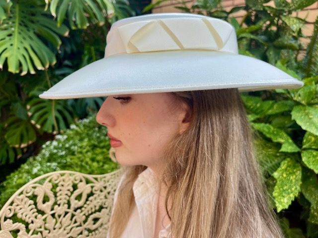 Woman wearing a white wide-brimmed hat with light-yellow accents, outdoors with green foliage.