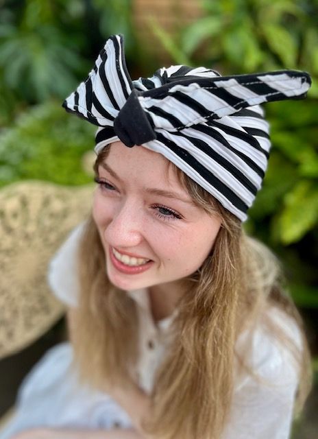 Woman wearing a black and white striped turban, smiling outdoors, surrounded by greenery.