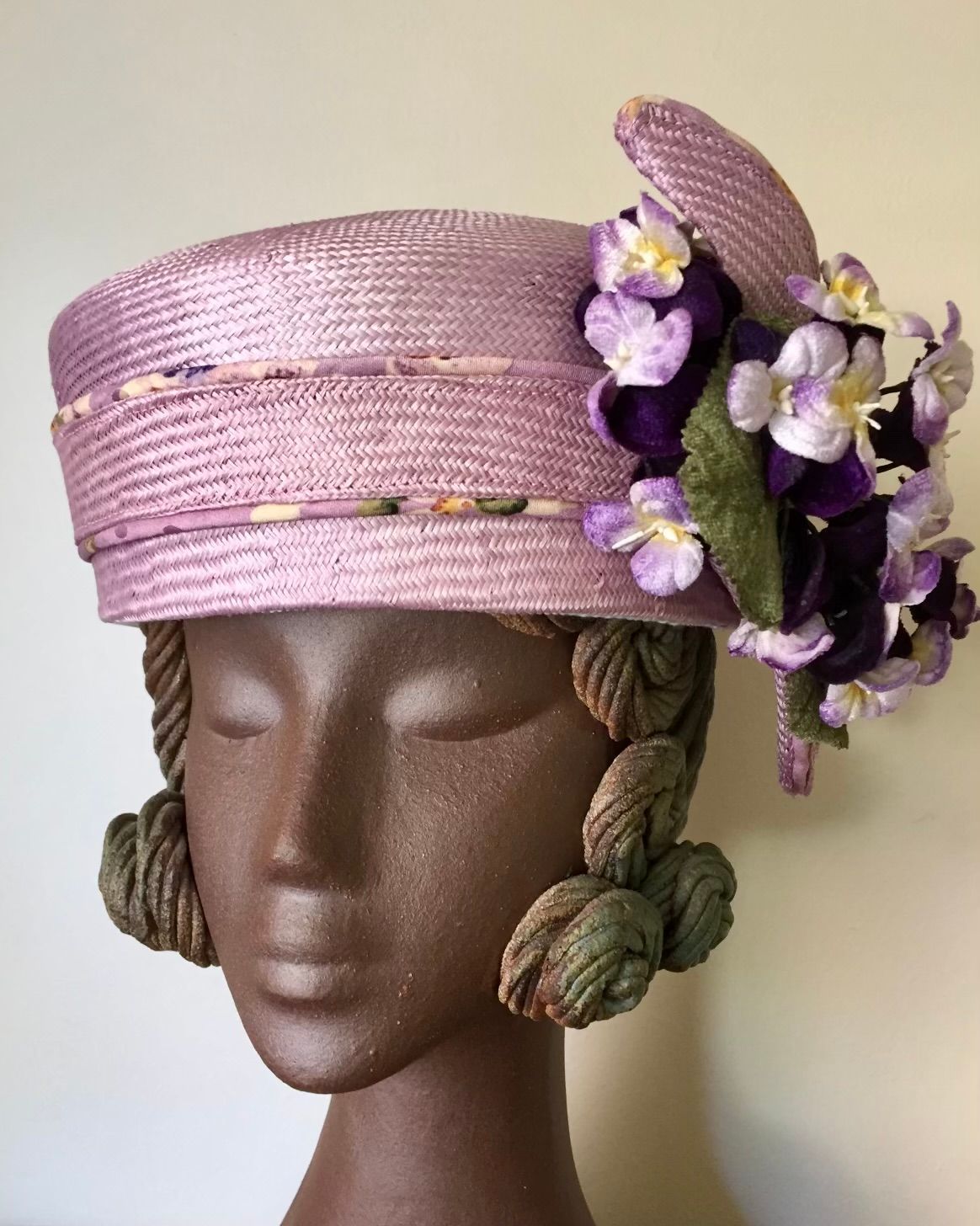 A lilac straw hat with floral accents sits on a mannequin head with decorative hair.