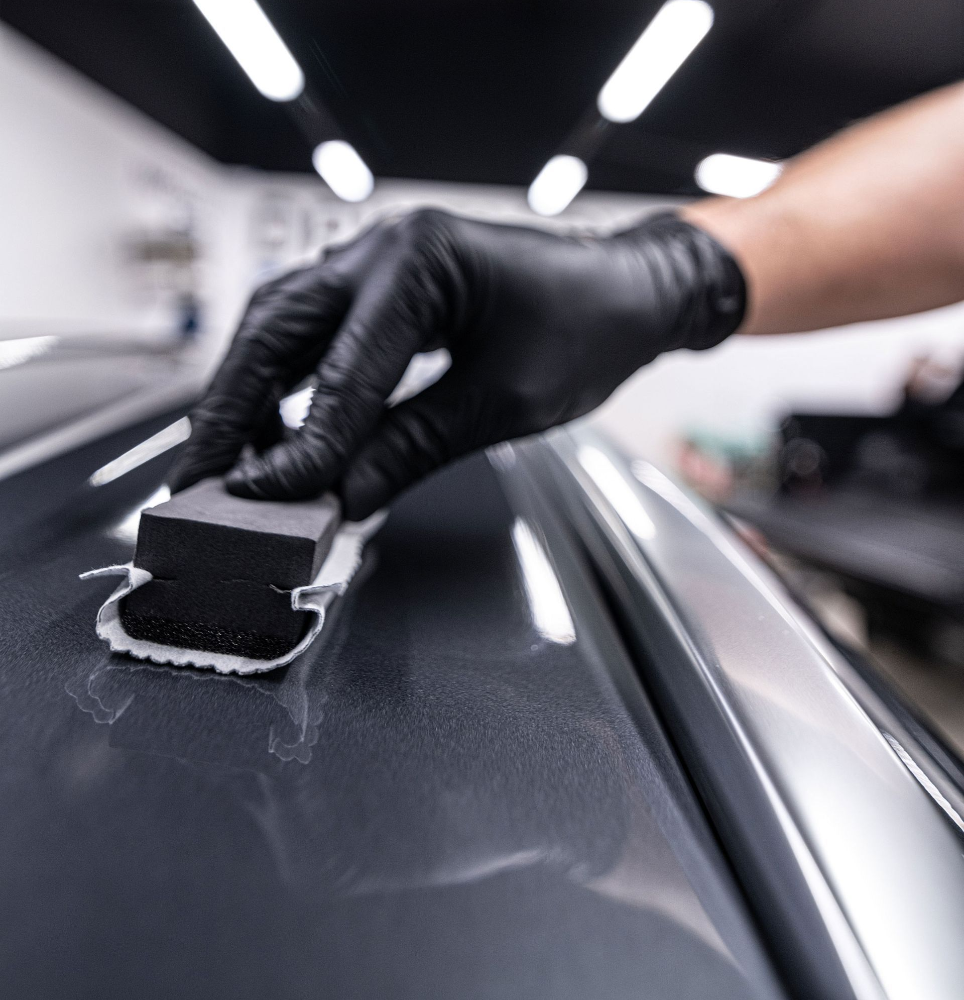 Gloved hand wiping a dark car surface with a sponge in a detailing shop