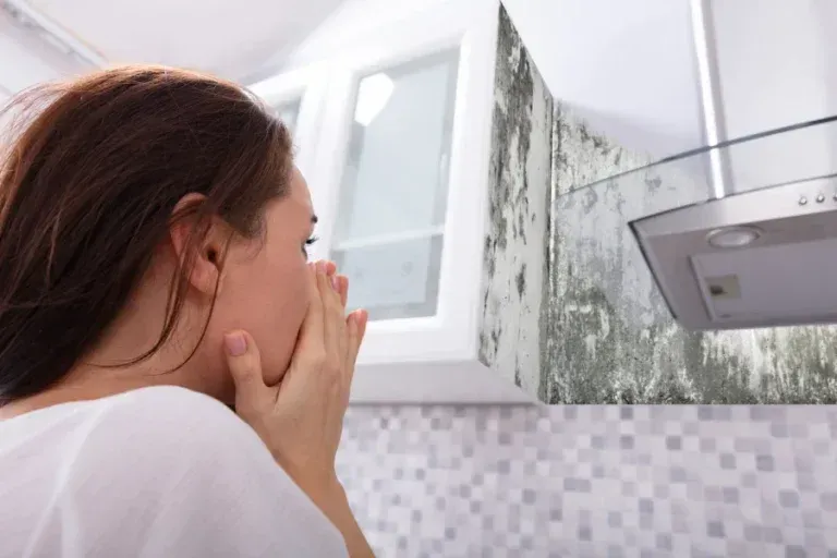 A person looks shocked at a kitchen wall covered in dark, extensive mold growth near white cabinets and a vent hood.